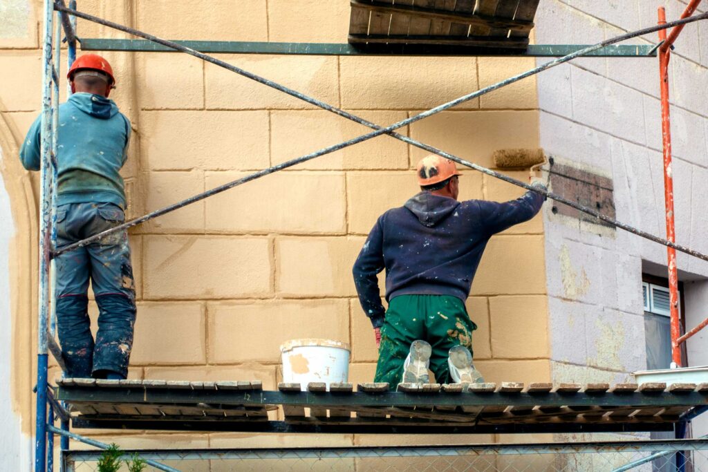Commercial painting contractors standing on a scaffolding painting a brick wall a cream color